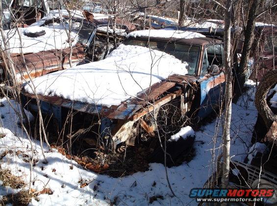 Snow-covered, rusty car with blue and brown paint, front end damaged, in a wooded area.