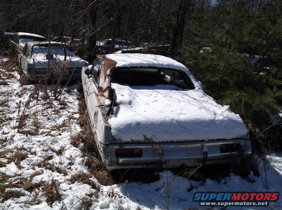 Two old cars sit in a snowy junkyard, one with its roof caved in