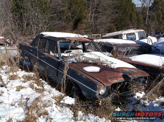 Rusty dark blue sedan in a snowy junkyard, with visible damage and vegetation.