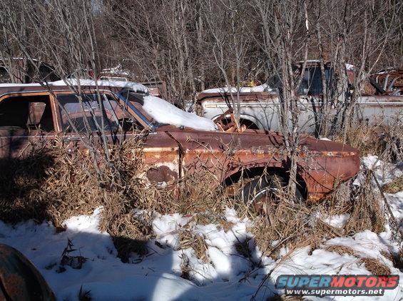Rusted vintage car in a snowy junkyard with a truck behind it.