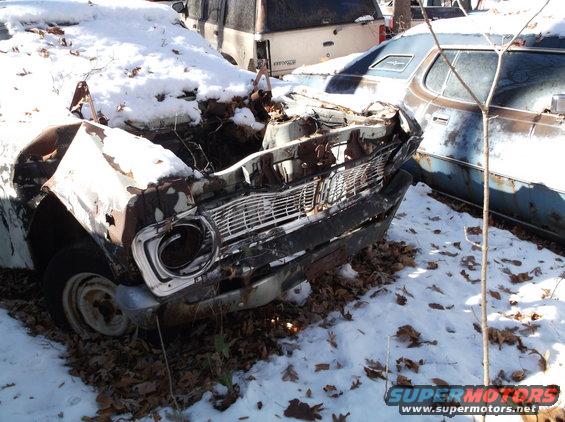 Front end of a heavily damaged car, possibly a 1970s model, covered in snow. Another car is visible to the right