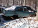 1974 Dodge Challenger - An abandoned aqua sedan sits in a snowy junkyard, missing its wheels and showing signs of rust.