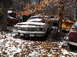 1974 Dodge Challenger - An old sedan sits in a snowy junkyard, surrounded by trees and fallen leaves.