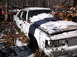 1974 Dodge Challenger - A white sedan sits abandoned in a snowy junkyard, partially covered by snow and fallen leaves.