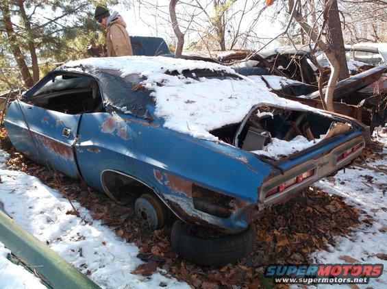 Abandoned blue 1974 Dodge Challenger coupe in snow, missing wheels and showing rust.