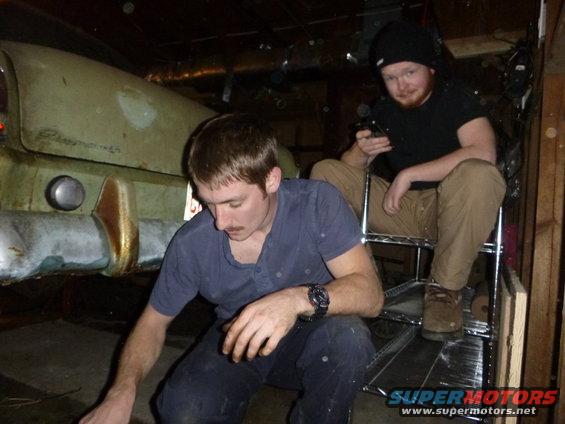 guy working on rear of old car, another guy sitting on shelf watching phone
