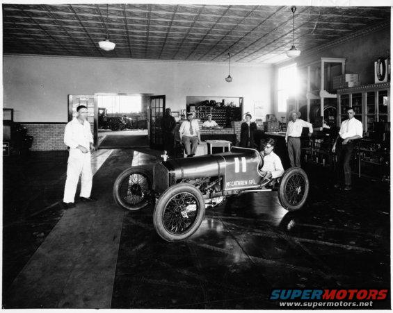 Racing car with number 11 and "MCCATHREN SPL" text on its side, parked indoors with several people around