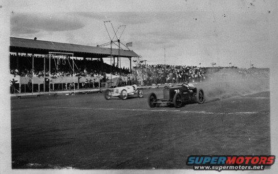 Two race cars compete on a track in front of a grandstand full of spectators. One car kicks up dust