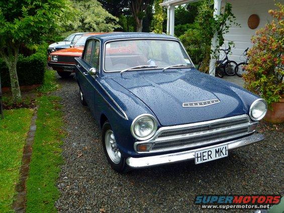 Blue sedan with 'Cortina' badge on hood, parked in gravel driveway next to an orange SUV and a white house