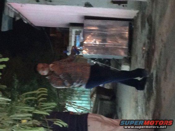 Person in plaid shirt and dark pants standing outdoors at night near a metal structure and plants