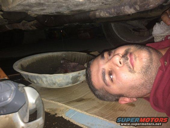 Man lying on the ground under a vehicle, with an oil drain pan and a jug of oil nearby