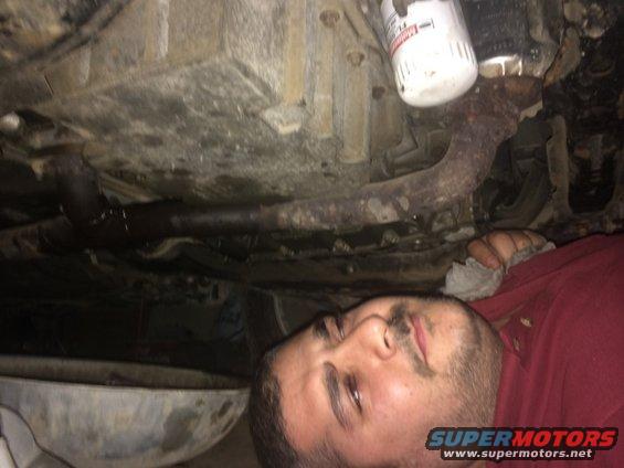 Underneath a vehicle, showing the exhaust pipe, oil filter, and part of the engine block. A person is lying down, looking at the components