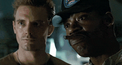 Two men in a dimly lit scene, one looking towards the camera
