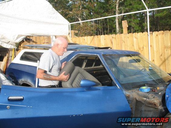 man holding car seat, blue car in foreground, blue and white SUV behind it