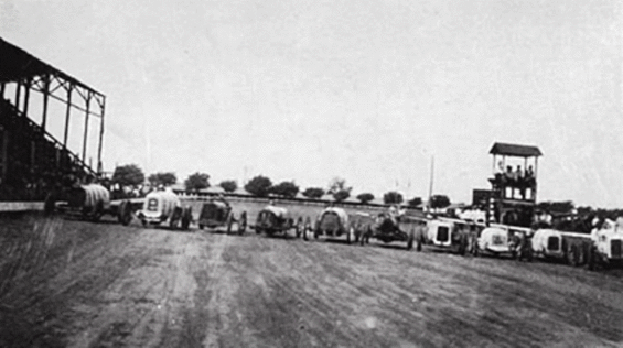 Multiple early 20th-century automobiles lined up on a track with a grandstand in the background