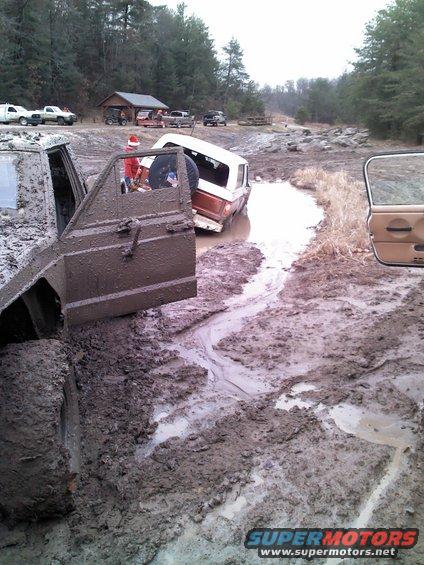 Mud-covered 1987 Jeep Comanche pickup truck with door open, stuck in a muddy ditch