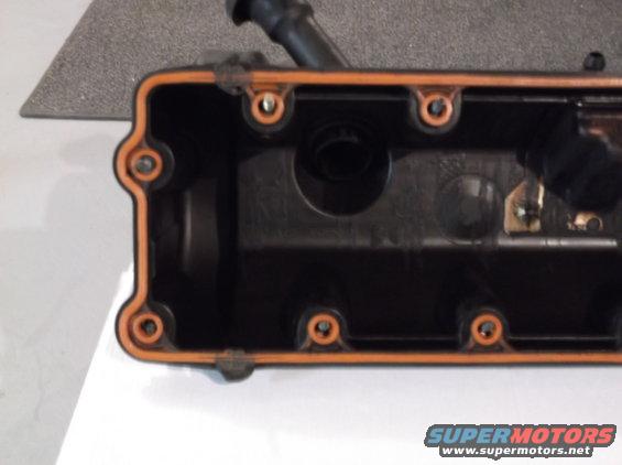 Close-up of an engine valve cover with an orange gasket. Some bolts are visible