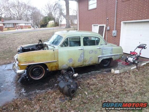 Old green sedan with hood open, engine removed, parked in driveway next to a garage