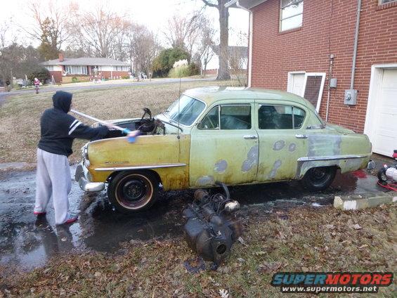 Green sedan undergoing restoration, hood open, engine removed, person washing front fender
