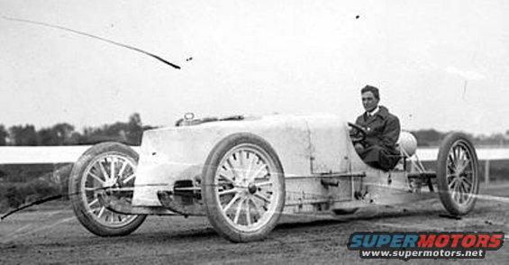 Driver in a vintage race car with spoked wheels and a tall radiator shroud