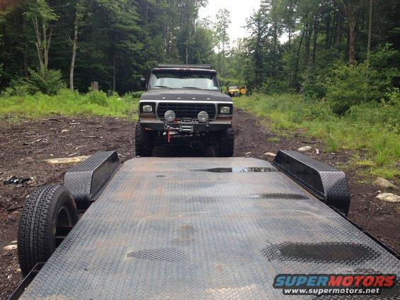 Ford Bronco with winch and lights on a dirt road, trailer in foreground. Yellow vehicle in background