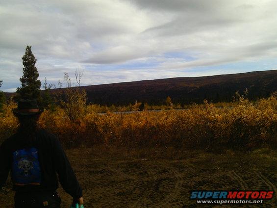 Person wearing a hat looks out at a landscape with dry grass and trees under a cloudy sky. A watermark reads SUPERMOTORS