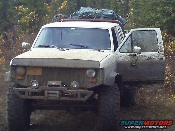 White pickup truck with off-road modifications, mud splattered, and gear on the roof rack