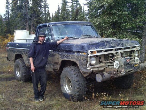 Mud-covered dark blue Ford truck, 1970s model, with a man standing beside it. Winch mounted on front bumper