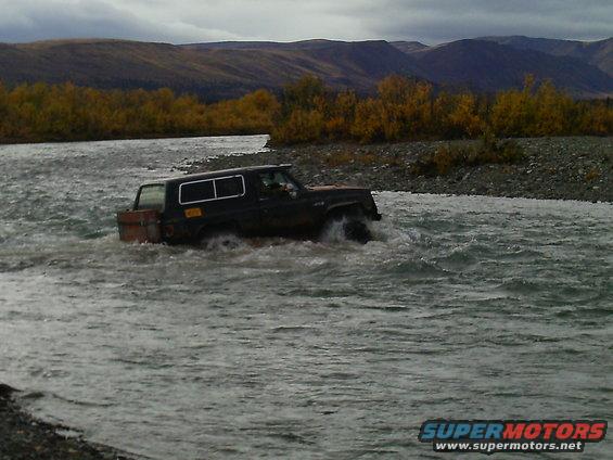 Black SUV driving through a river with trees and mountains in the background