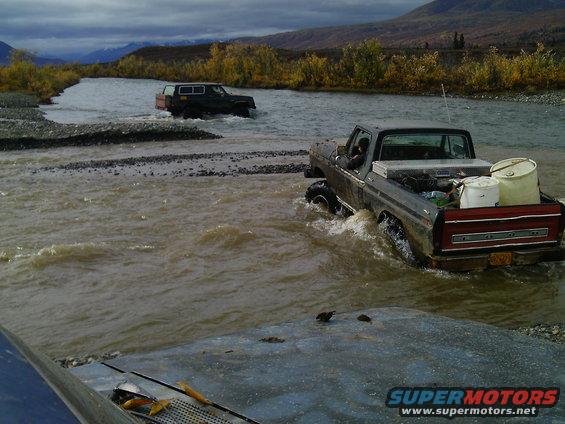 Gray Ford pickup truck with red trim driving through river, carrying supplies. Another SUV is in the background