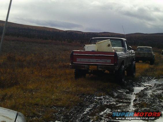 Rear view of a red 1979 Ford F-150 pickup truck loaded with white containers, parked in a muddy field