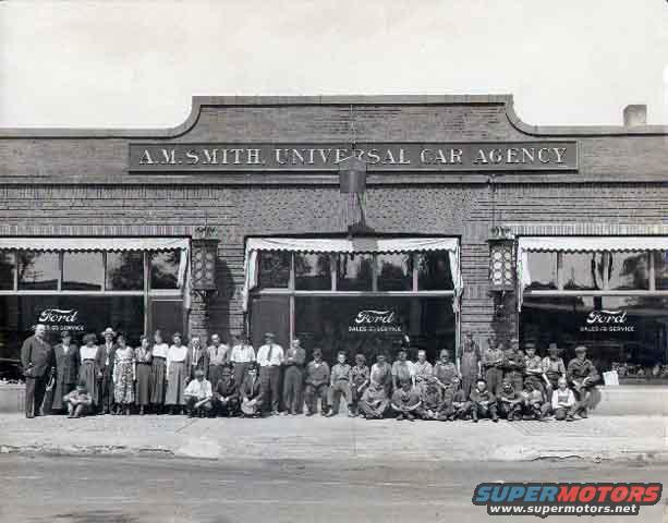 Group of people standing and sitting in front of A.M. Smith Universal Car Agency building with Ford signs