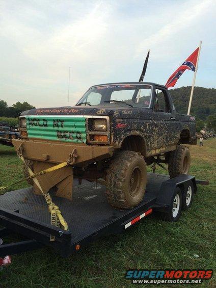 Mud-covered Ford Bronco on a trailer, green painted grille with text, Confederate flag on antenna