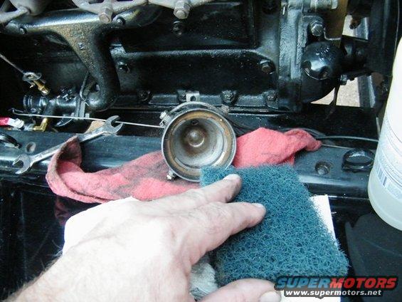 Hand cleaning a metal engine part with a blue scrub pad next to a red rag and wrenches