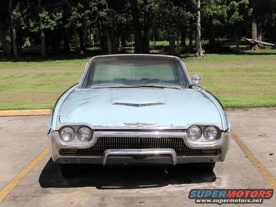 Front view of a light blue 1963 Ford Thunderbird parked in a lot with trees in the background