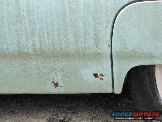 Light blue car body panel with rust spots and scratches, showing a tire and part of a wheel well