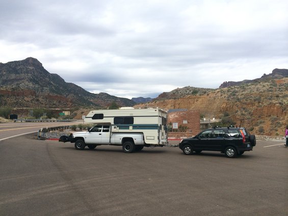 White pickup truck with camper shell towing a black SUV on a road with mountains in the background