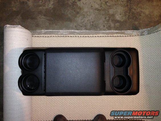 Black center console with four cup holders and a textured rectangular panel, resting on a carpeted surface