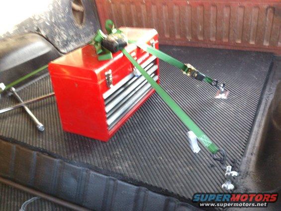 Red toolbox secured with green straps in a truck bed with a spare tire nearby