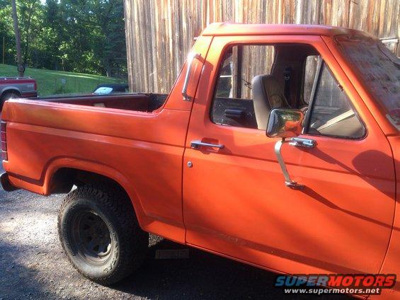 Orange SUV, side view, with chrome side mirror and black tire. Another red vehicle is visible in the background