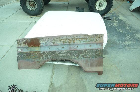 Rusted quarter panel on top of a white hood, with large tires in the background