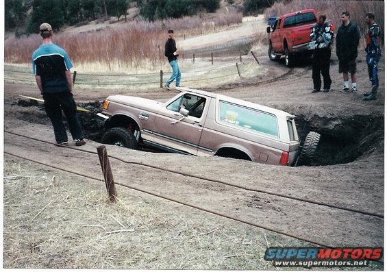 1996 Ford Bronco stuck in a deep dirt hole with a red pickup truck nearby