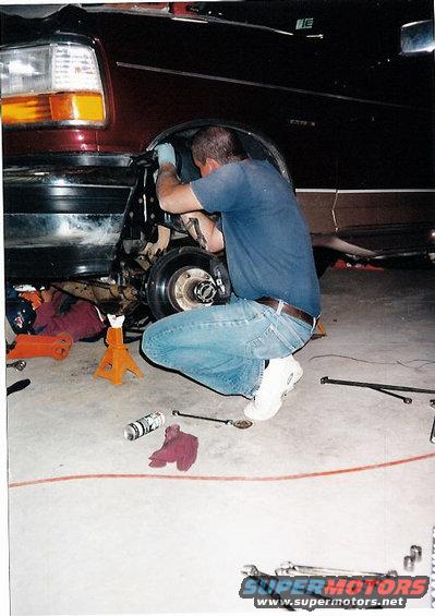 Maroon SUV with front fender removed, man working on suspension. Tools and jack stands on floor