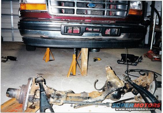 Maroon Ford truck front end on jack stands, with disassembled front axle on the floor