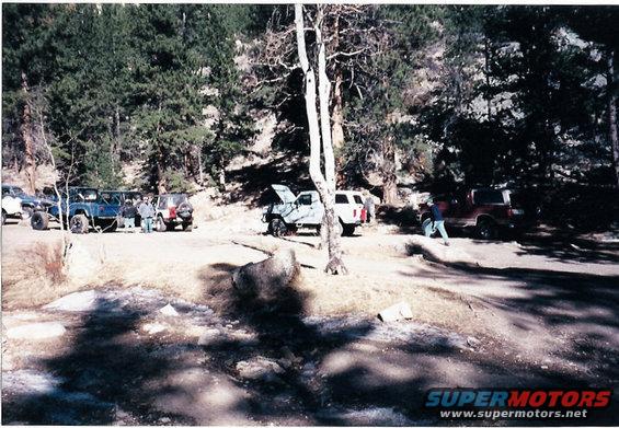 Several SUVs and off-road vehicles parked on a dirt lot in a forest. One vehicle has its hood open