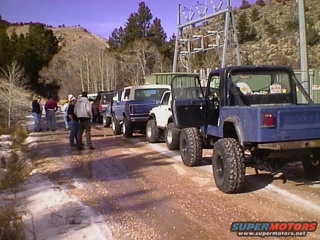 Line of trucks and SUVs parked on a dirt road with snow, mountains in background