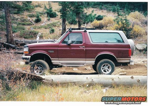 Maroon Ford Bronco with a white roof and spare tire on the back, parked on a dirt road with trees and brush