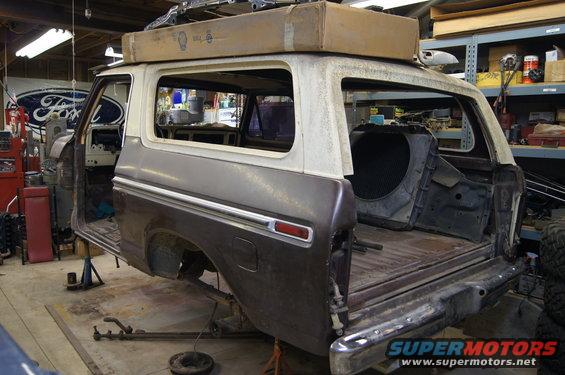 Ford Bronco body on jack stands in a garage, stripped down for repair. A large box is on the roof