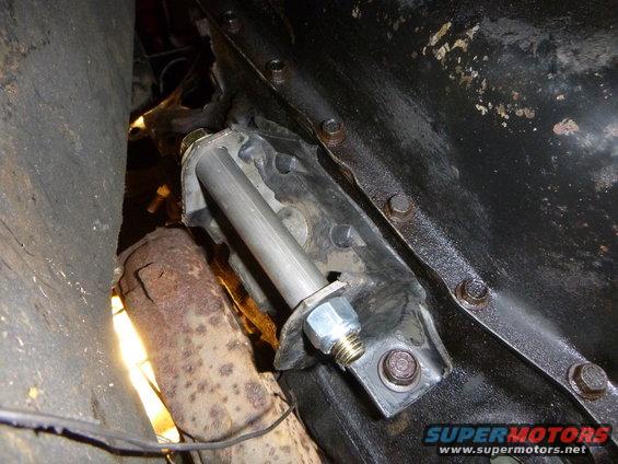 Close-up of a metal bracket bolted to a dark engine component, with a rusty exhaust pipe nearby