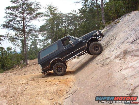 Black Ford Bronco climbing a steep rock face, side view, with trees in background